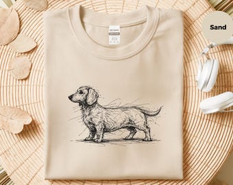 May include: A sand-colored crew neck t-shirt featuring a black line-art sketch of a dachshund dog on the front. The shirt has a Gildan brand label at the inner neckline and a circular tag in the upper right corner with the text Sand.