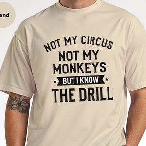 May include: A sand-colored short-sleeve t-shirt featuring black text that reads: NOT MY CIRCUS NOT MY MONKEYS BUT I KNOW THE DRILL. The design includes two small stars flanking the phrase BUT I KNOW on a horizontal banner graphic.
