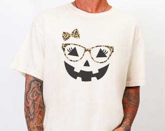May include: A cream-colored short-sleeve t-shirt featuring a graphic of a jack-o'-lantern face. The pumpkin has black triangular eyes with eyelashes, a black jagged mouth, and wears leopard print glasses and a matching leopard print bow.