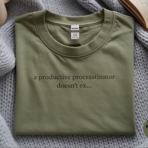 May include: A military green crew neck t-shirt featuring the black printed text 'a productive procrastinator doesn't ex...' centered on the chest. The shirt has a standard collar with a white brand label visible at the inner neckline.