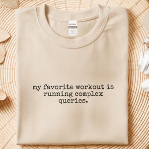 Può includere: T-shirt color sabbia con la scritta nera "my favorite workout is running complex queries". La maglietta è piegata su una superficie intrecciata, con cuffie bianche nelle vicinanze. La maglietta è del marchio Gildan.