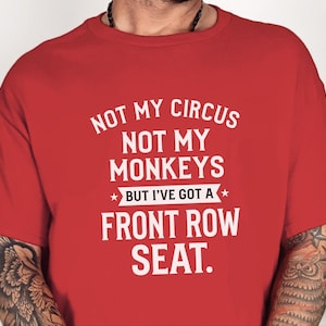 May include: A red short-sleeved t-shirt featuring white text that reads: NOT MY CIRCUS NOT MY MONKEYS BUT I'VE GOT A FRONT ROW SEAT. The shirt has a crew neckline and is made of a solid-colored fabric with a screen-printed graphic on the front.