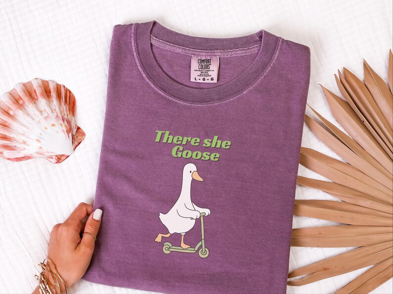 May include: A plum-colored t-shirt featuring a graphic of a white goose riding a light green scooter. Above the goose, the text reads "There she Goose" in light green lettering. The shirt has a crew neckline and a Comfort Colors brand label.