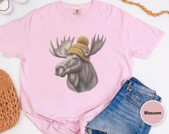 May include: A light pink short-sleeve t-shirt featuring a graphic of a grey moose wearing a mustard yellow knit beanie with a pom-pom. The shirt has a crew neckline and a small brand label printed on the inside of the collar area.