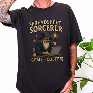 May include: A black t-shirt featuring a graphic of a wizard working on a laptop with a staff and a cup of coffee. The text above reads SPREADSHEET SORCERER and the text below reads SUM (= COFFEE). The design includes stars and a glowing orb.