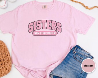 May include: A light pink short-sleeve t-shirt featuring the text "SISTERS" in a large, arched, black-outlined pink font, with "FROM OTHER MISTERS" printed in a smaller, rectangular, pink-filled box with black outlines.