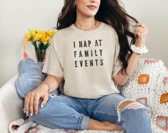 May include: A beige short-sleeved t-shirt featuring the black text I NAP AT FAMILY EVENTS printed in a bold, sans-serif font across the chest. The shirt is paired with blue denim jeans that have distressed rips at the knees.