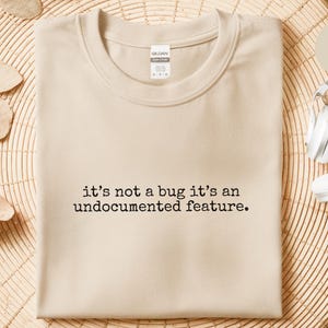 Peut inclure: T-shirt beige avec l'inscription noire : "it's not a bug it's an undocumented feature". Le t-shirt est plié sur un support tressé, avec un casque blanc à proximité. Le t-shirt est de couleur neutre.