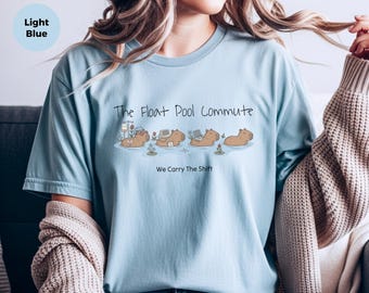 May include: A light blue t-shirt featuring a graphic of four capybaras swimming in water. The text reads The Float Pool Commute and We Carry The Shift. The capybaras are depicted with various items including an IV pole, a laptop, and a bag.