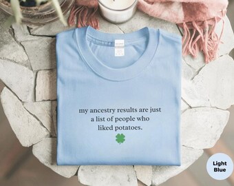 May include: A light blue t-shirt featuring the black text "my ancestry results are just a list of people who liked potatoes." and a small green four-leaf clover icon centered below the text. The shirt has a crew neckline and a white tag.