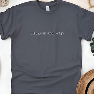 Puede incluir: Una camiseta gris oscuro con el texto "git push and pray" en blanco, con una fuente sencilla. La camiseta tiene las mangas enrolladas y cuello redondo. La camiseta se muestra sobre una superficie blanca.