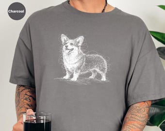 May include: A charcoal gray short-sleeve t-shirt featuring a white line-art sketch of a Corgi dog standing in profile. The graphic is centered on the chest and depicts the dog with its mouth open. The shirt has a crew neckline and a relaxed fit.