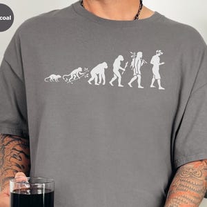 Può includere: Una t-shirt grigio antracite a maniche corte con una stampa grafica bianca che raffigura l'evoluzione da un piccolo mammifero a un essere umano moderno che tiene in mano uno smartphone. Il disegno è centrato sul petto della maglia a girocollo.