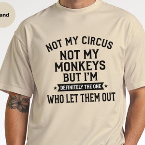 May include: A sand-colored short-sleeve t-shirt featuring black text that reads: NOT MY CIRCUS NOT MY MONKEYS BUT I'M DEFINITELY THE ONE WHO LET THEM OUT. The design includes a horizontal banner element with two small stars.