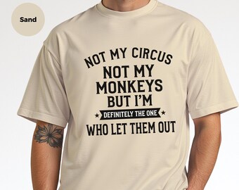 May include: A sand-colored short-sleeve t-shirt featuring black text that reads: NOT MY CIRCUS NOT MY MONKEYS BUT I'M DEFINITELY THE ONE WHO LET THEM OUT. The design includes a horizontal banner element with two small stars.