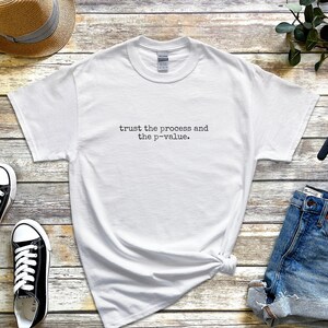 Könnte beinhalten: Weißes T-Shirt mit dem Text "trust the process and the p-value." Das Bild zeigt auch blaue Jeansshorts, schwarze Turnschuhe, einen Strohhut, eine Smartwatch und kabellose Ohrhörer. Die Artikel sind auf einer Holzoberfläche angeordnet.