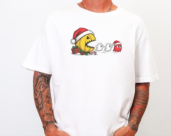 May include: A white t-shirt featuring a graphic of a yellow, round, mouth-open character wearing a Santa hat, chasing two white ghosts and one red ghost wearing a Santa hat, with a wrapped gift and a candy cane at the base.