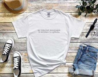 Puede incluir: Una camiseta blanca de manga corta con el texto negro en estilo de máquina de escribir "my internal monologue is written in python." centrado en el pecho. La prenda tiene un cuello redondo clásico y un corte estándar, presentada como un artículo plano.
