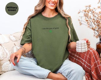 May include: A military green short-sleeve t-shirt featuring the black text "i am the pot of gold." with a small green circle icon below the lettering. The shirt is worn with light blue denim jeans and paired with a white and brown grid mug.