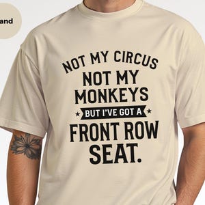 May include: A sand-colored short-sleeve t-shirt featuring black text that reads: NOT MY CIRCUS NOT MY MONKEYS BUT I'VE GOT A FRONT ROW SEAT. The design includes a banner graphic with two small stars on either side of the phrase BUT I'VE GOT A.