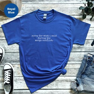 Puede incluir: Camiseta azul real con el texto "sorry for what I said during the merge conflict." La camiseta está combinada con zapatillas deportivas blancas y negras, pantalones cortos vaqueros, un reloj y un sombrero.