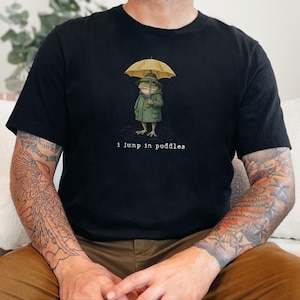 May include: A black short-sleeve t-shirt featuring a graphic of a frog wearing a green raincoat and hat while holding a yellow umbrella. Below the frog, the text reads "I jump in puddles" in a typewriter-style font.