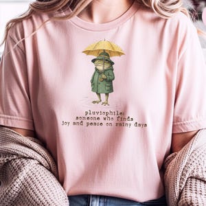 May include: A light pink t-shirt featuring a graphic of a frog wearing a green raincoat and holding a yellow umbrella. Below the frog, the text reads: pluviophile: someone who finds joy and peace on rainy days. The shirt has short sleeves.