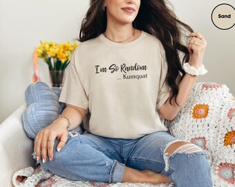 May include: A sand-colored short-sleeve t-shirt featuring the black printed text "I'm So Random ... Kumquat" centered on the chest. The shirt has a crew neckline and a relaxed fit, constructed from a solid light beige fabric.