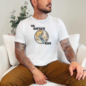 May include: A white short-sleeved t-shirt featuring a circular graphic on the chest. The graphic depicts a comic-style illustration of a person in a kitchen with the text YA SISTA'S ASS printed in bold black lettering around the circular design.