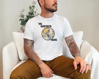 May include: A white short-sleeved t-shirt featuring a circular graphic on the chest. The graphic depicts a comic-style illustration of a person in a kitchen with the text YA SISTA'S ASS printed in bold black lettering around the circular design.