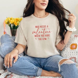 May include: A sand-colored short-sleeve t-shirt featuring the red text Who Needs A Valentine When You Have The Gym with two small heart icons. The shirt is paired with blue denim jeans that have distressed detailing at the knees.
