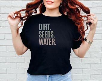 Könnte beinhalten: Ein schwarzes kurzärmeliges T-Shirt mit dem Text DIRT. SEEDS. WATER. in einer gestapelten, serifenlosen Schrift auf der Brust. Das Wort DIRT ist hellgrau, SEEDS ist in einem gedämpften Salbeigrün und WATER in einem sanften Hellbraun gehalten.