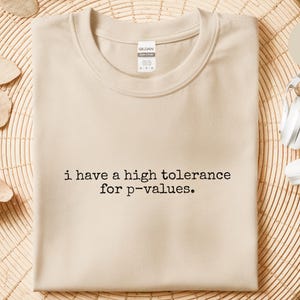 May include: A sand-coloured crew neck t-shirt featuring the black text "i have a high tolerance for p-values." printed in a typewriter-style font across the chest. The shirt has a Gildan brand label visible at the inner neckline.
