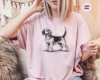 May include: A light pink t-shirt featuring a black and white sketch-style illustration of a beagle dog standing in profile. The dog is depicted with detailed line work, and the shirt includes a small circular tag labeled Light Pink.
