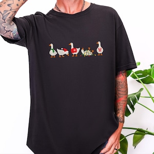 May include: A black short-sleeved t-shirt featuring a graphic print of five white geese in a row. The geese are wearing various holiday-themed accessories including a green scarf, a red Santa hat, a red sweater, tangled string lights, and a red gift box.
