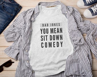 May include: A light gray short-sleeved t-shirt featuring the black text DAD JOKES YOU MEAN SIT DOWN COMEDY. The shirt is layered under a white and gray striped button-down shirt, accompanied by blue jeans, dark sunglasses, and blue sneakers.