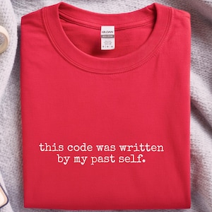 May include: A bright red t-shirt with the text "this code was written by my past self" in white. The shirt is folded neatly on a gray blanket, with a coffee mug and notebook visible in the background. The shirt is a Gildan brand.