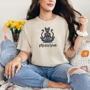 May include: A beige short-sleeve t-shirt featuring a graphic of a black cat sitting in a lotus yoga position with colorful geometric patterns. Below the cat, the text Meowisim is printed in a black cursive font. The shirt has a crew neckline.