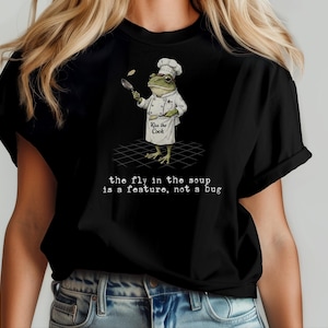 Könnte beinhalten: Ein schwarzes T-Shirt mit einer Grafik eines Frosches, der eine weiße Kochmütze und eine Schürze mit der Aufschrift Kiss the Cook trägt. Der Frosch hält eine Pfanne und einen Pfannenwender. Darunter steht der Text, the fly in the soup is a feature, not a bug.
