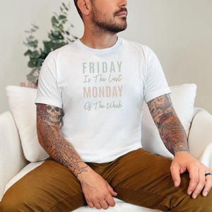 Può includere: Una t-shirt bianca a maniche corte con la scritta "FRIDAY Is The Last MONDAY Of The Week" stampata sul petto. La parola "FRIDAY" è verde salvia, "MONDAY" è rosa tenue e le restanti parole sono grigio scuro.