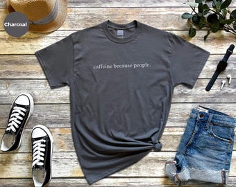 Op de afbeelding: Een antracietgrijs T-shirt met korte mouwen en een ronde hals met de witte tekst "caffeine because people." op de borst gedrukt. Het shirt wordt getoond naast een strooien hoed, zwarte sneakers, een blauwe spijkerbroekshort en een zwart horloge.