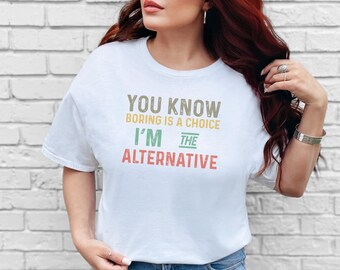 May include: A white short-sleeve t-shirt featuring the text "YOU KNOW BORING IS A CHOICE I'M THE ALTERNATIVE" printed in a multi-colored, distressed font. The words are stacked in shades of grey, yellow, green, and red.