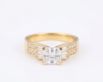 Pode incluir: Um anel de ouro com uma pedra preciosa central transparente de corte quadrado, ladeada por pedras de corte baguette em cada lado. A banda é decorada com duas filas de pequenas pedras redondas transparentes ao longo das laterais do anel.