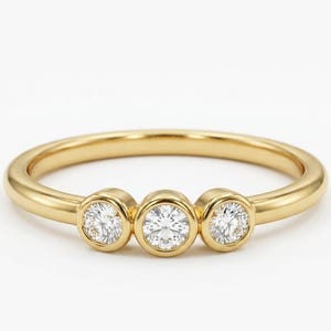 May include: A gold ring with three round, clear gemstones set in individual bezels. The band is a smooth, polished gold. The gemstones are evenly spaced across the top of the ring.