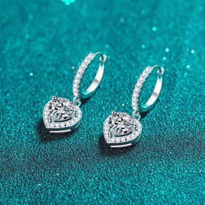 May include: A pair of silver hoop earrings with heart-shaped pendants. Each pendant features a central, clear gemstone surrounded by smaller stones. The hoops are also adorned with small, clear stones, creating a sparkling effect. The earrings are displayed on a teal surface.