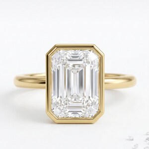 May include: A gold ring featuring a large, rectangular, emerald-cut diamond. The diamond is set in a bezel setting, and the band is a simple, polished gold. The ring is displayed on a white background.