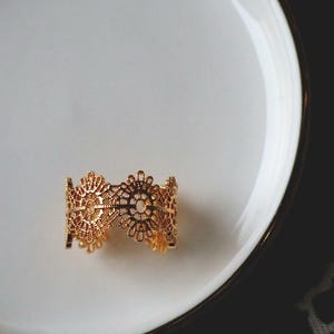 Può includere: Un bracciale a polsino traforato color oro con un motivo floreale poggia su un piatto bianco. Il bracciale presenta motivi simmetrici intricati e dettagli delicati. Il piatto ha un bordo dorato.