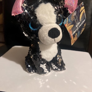 May include: A black and white plush dog with pink ears and blue eyes. The dog is covered in sequins and has a tag that says "6.5in Forest Friend".