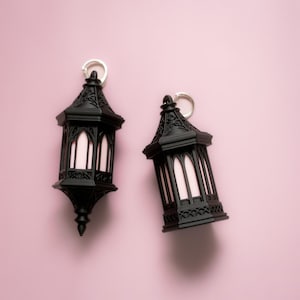 Puede incluir: Dos llaveros de linterna negra en miniatura con detalles ornamentados y un interior blanco, sobre una superficie rosa claro. Cada linterna tiene un llavero plateado en la parte superior.