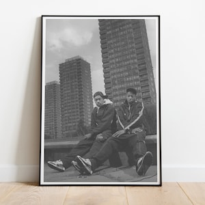 Può includere: Due persone con giacche e cappelli neri sono sedute su un cornicione di fronte a un alto edificio di appartamenti. L'edificio è in cemento e ha molte finestre. La foto è in bianco e nero.